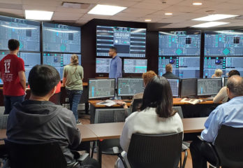 Nine people seated at tables or standing as they observe data on a wall of large computer monitors.